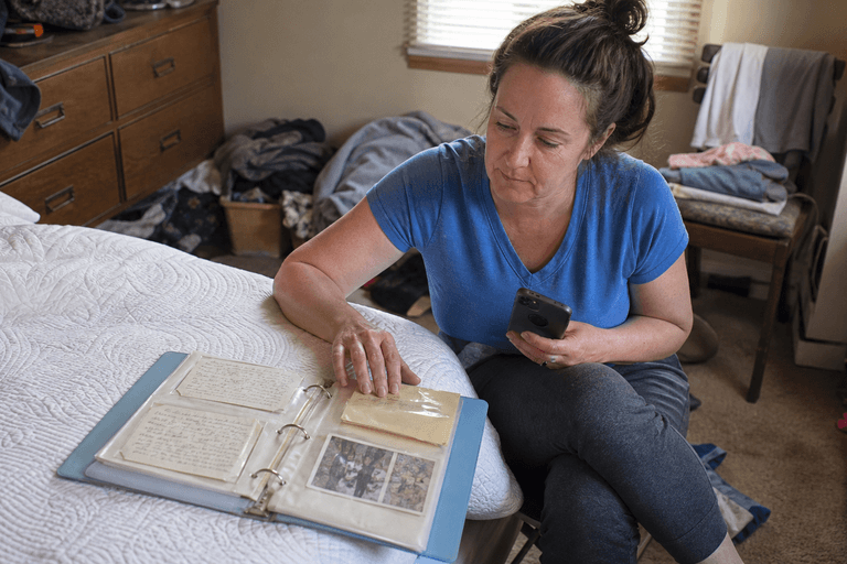 A woman sits beside a bed looking through a binder of her mother's recipe cards and photographs