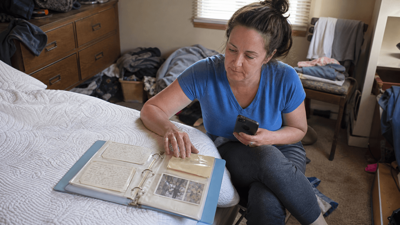 A woman sits beside a bed looking through a binder of her mother's recipe cards and photographs