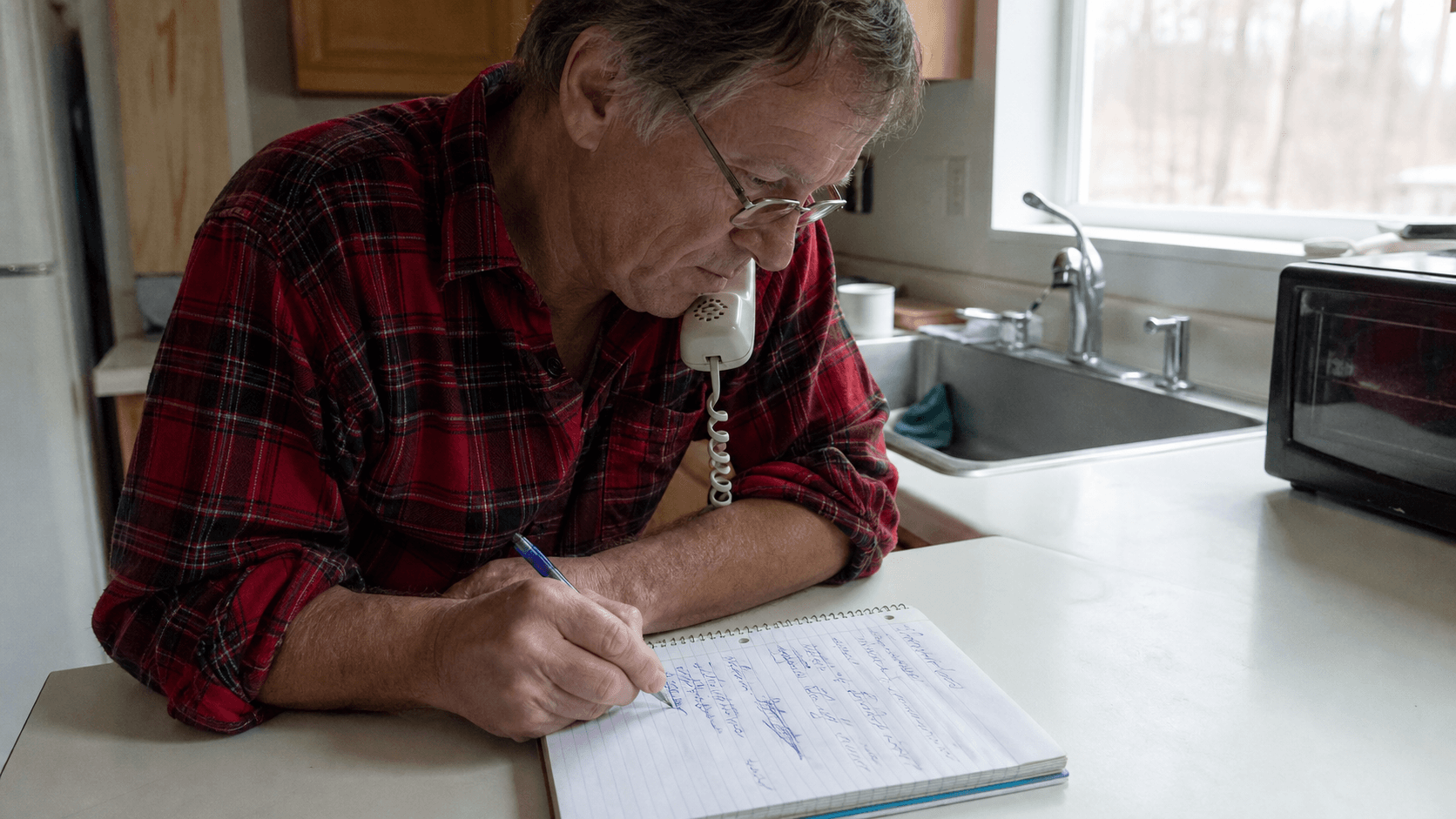 A man at a kitchen counter writes notes on a spiral notepad while on the phone